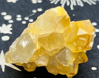 May include: A cluster of golden-yellow quartz crystals. The crystals are clustered together, with sharp, geometric facets. The background is a dark blue surface with white celestial designs and the text "ST1".