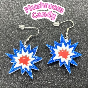 Puede incluir: Un par de pendientes de estrella azul, blanco y rojo con ganchos plateados. Los pendientes están hechos de purpurina y tienen un efecto de capas.