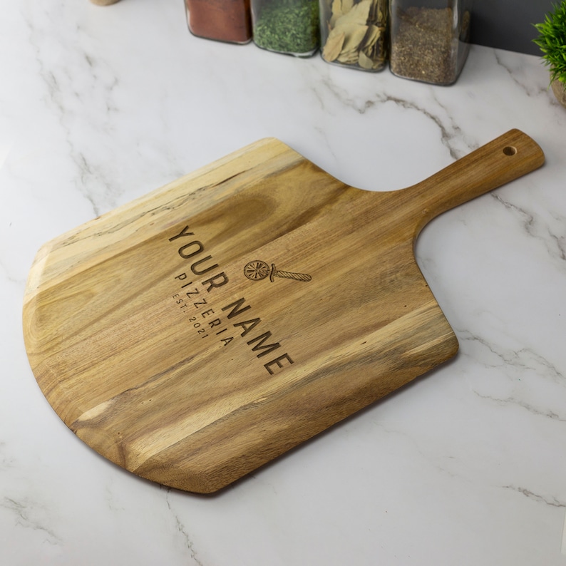 May include: A wooden pizza peel with a handle. The peel is engraved with the text "YOUR NAME PIZZERIA EST: 2021" and a pizza slice icon.