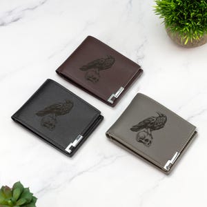 Pode incluir: Três carteiras de couro em preto, marrom e cinza, cada uma com um corvo empoleirado em um crânio. As carteiras têm um design bi-fold com um detalhe de canto de metal prateado. Fundo de mármore branco.