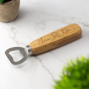 May include: A wooden bottle opener with a metal opener and a handle engraved with "Chris & Rita Est. 2025" and a house icon.