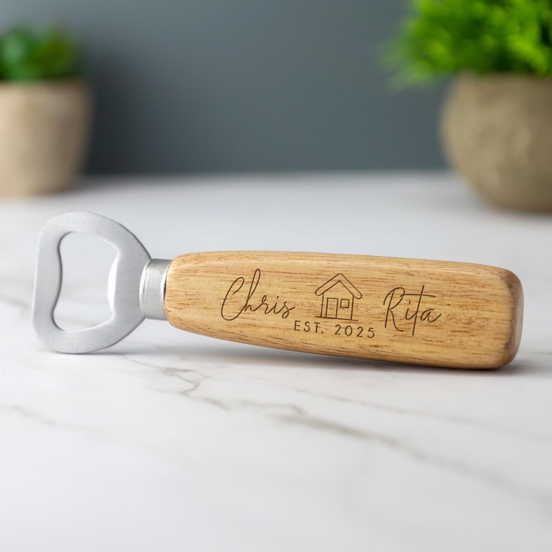 May include: A wooden bottle opener with a metal cap opener. The handle is engraved with the names "Chris" and "Rita", a house icon, and the text "EST. 2025".
