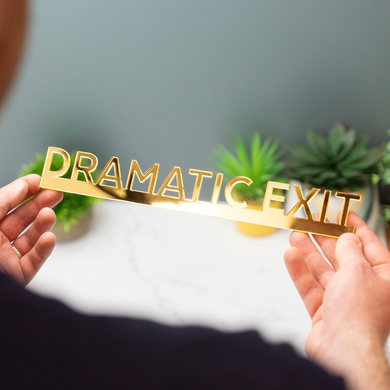 K&ouml;nnte beinhalten: Ein goldfarbenes Metallschild mit den ausgeschnittenen Worten "Dramatic Exit". Das Schild wird von den H&auml;nden einer Person gehalten.