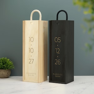 May include: Two wooden wine boxes with engraved dates and names. The box on the left is light wood with a white rope handle and says "ENGAGED Reece & Keni 10.10.27". The box on the right is dark wood with a black rope handle and says "MARRIED Vick & Nick 05.12.26".