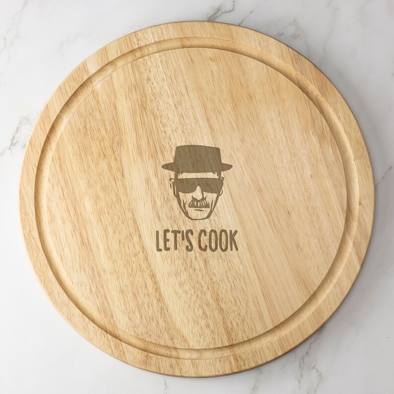 May include: A round wooden cutting board with a dark brown rim and a laser-engraved image of a man wearing a hat and sunglasses. The text "LET'S COOK" is engraved below the image.
