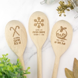 May include: Three wooden spoons with Christmas-themed designs and text. The first spoon has a candy cane design and the text "Kiera Cane Do It". The second spoon has a snowflake design and the text "Love at Frost-Sight Megan & Joe". The third spoon has a Christmas pudding design and the text "All Good In The Pud".