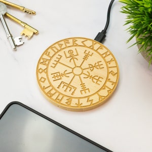 May include: A wooden wireless charging pad with a circular design featuring a compass rose and runes around the edge. The charging pad is connected to a USB cable.