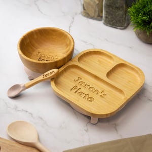 May include: A wooden baby feeding set with the name "Jaxon" engraved on the bowl, plate, and spoon. The set includes a divided plate, a bowl, and a spoon with a pink silicone tip. The plate and bowl are on a light pink base.
