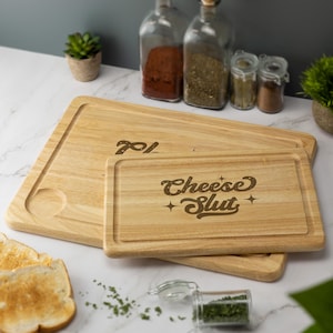 May include: Two wooden chopping boards, one larger and one smaller, stacked. The smaller board has the text "Cheese Slut" engraved. The larger board has the number "701" engraved. The boards are light brown in colour.
