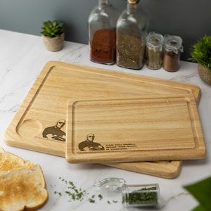May include: Two wooden chopping boards with a  "Can you smell what The Rock is cooking?" inscription and a silhouette of Dwayne Johnson. The smaller board is on top of the larger board.