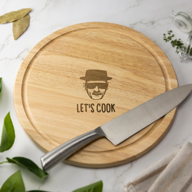 May include: A round wooden cutting board with a laser-engraved image of a man wearing a hat and sunglasses. The text "LET'S COOK" is engraved below the image. A silver knife is lying on the board.