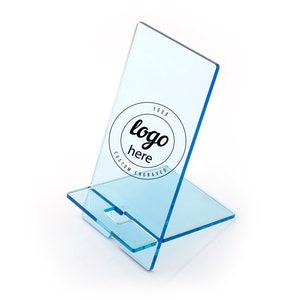 May include: A clear blue acrylic phone stand with a slot for a phone. The stand has a circle engraved with the text "YOUR logo here CUSTOM ENGRAVED".