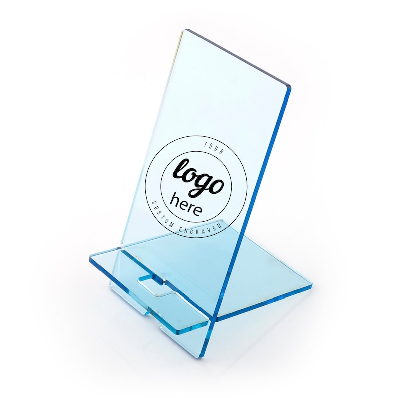 May include: A clear blue acrylic phone stand with a slot for a phone. The stand has a circle engraved with the text "YOUR logo here CUSTOM ENGRAVED".