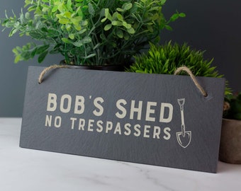 Puede incluir: Un letrero de pizarra negra con texto blanco que dice "Bob's Shed No Trespassers" con un pequeño gráfico de pala. El letrero está colgado de una cuerda y está delante de algunas plantas verdes.