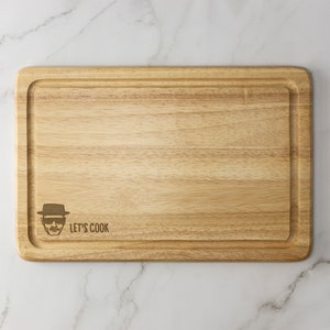 May include: A wooden cutting board with a light brown finish. It has a raised edge and a groove around the perimeter. The board has a laser-engraved image of a man wearing a hat and sunglasses with the text "Let's Cook" below it.