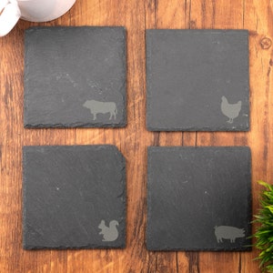 Puede incluir: Cuatro posavasos de pizarra negra con diferentes siluetas de animales. Los posavasos representan una vaca, una gallina, una ardilla y un cerdo.