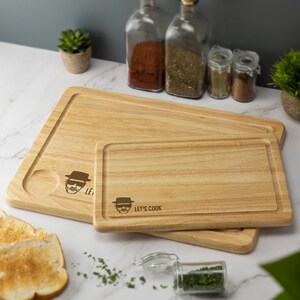 May include: Two wooden cutting boards with a light wood finish. The smaller board has a cartoon image of a man with the text "Let's Cook". The larger board has a cartoon image of a man with the text "Let's".