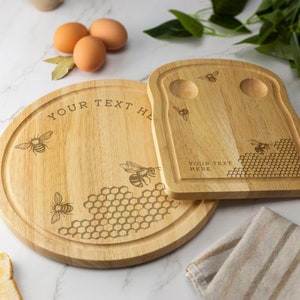 May include: Two wooden serving boards featuring a honeycomb and bee design. The larger board is round and has the text "YOUR TEXT HERE" engraved on it. The smaller board is shaped like a slice of bread and has two indentations for eggs. Both boards have a bee and honeycomb design.