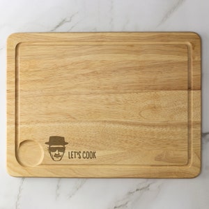 May include: A wooden cutting board with a rounded edge and a small juice groove. It features a laser-engraved image of a man wearing a hat and sunglasses with the text "Let's Cook".