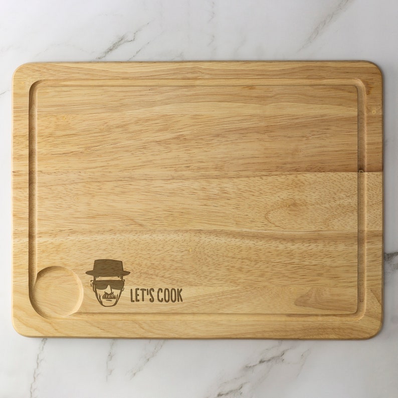 May include: A wooden cutting board with a rounded edge and a small juice groove. It features a laser-engraved image of a man wearing a hat and sunglasses with the text "Let's Cook".