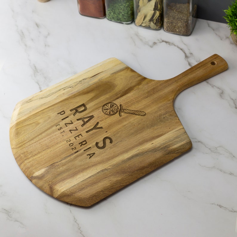 May include: A wooden pizza peel with a handle. The peel is engraved with the text "Ray's Pizzeria Est. 2021" and a logo of a pizza wheel with a knife.