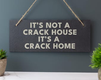 Puede incluir: Un letrero rectangular de pizarra negra con el texto "IT'S NOT A CRACK HOUSE IT'S A CRACK HOME" en letras blancas. El letrero está suspendido por una cuerda de hilo natural. El fondo es una pared de color verde azulado oscuro.