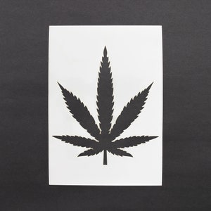 Könnte beinhalten: Eine schwarze Silhouette eines Cannabisblattes auf weißem, rechteckigem Papier. Die detaillierte Form des Blattes ist scharf gegen den weißen Hintergrund abgegrenzt. Das Papier liegt auf einem dunkelgrauen Hintergrund.