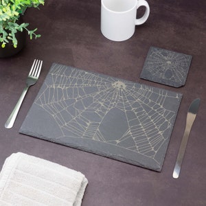 May include: A black slate coaster featuring a white spiderweb design. The coaster is rectangular with rounded corners. It is placed on a brown surface alongside a white mug and a silver fork.