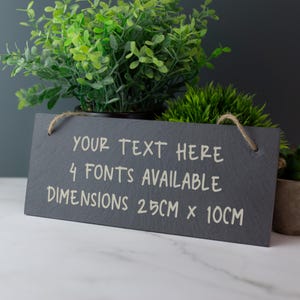Könnte beinhalten: Ein schwarzes Schiefer-Schild mit weißem Text, der "YOUR TEXT HERE 4 FONTS AVAILABLE DIMENSIONS 25CM x 10CM" lautet. Das Schild hängt an einem Stück Schnur.
