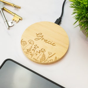 Könnte beinhalten: Ein rundes, drahtloses Ladegerät aus Holz mit einem floralen Design und dem eingravierten Namen "Gracie". Das Ladegerät ist mit einem schwarzen USB-Kabel verbunden.