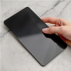 Puede incluir: Un objeto rectangular negro brillante con esquinas redondeadas. El objeto está sostenido por una mano y refleja la luz, creando reflejos. El fondo es una superficie clara y veteada. El objeto parece ser una pequeña tabla de cortar o una bandeja.