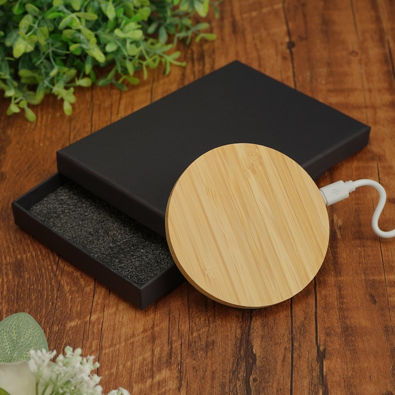 May include: A round, wooden wireless charging pad with a black fabric-lined box. The charging pad is made of light brown wood with a natural grain pattern.