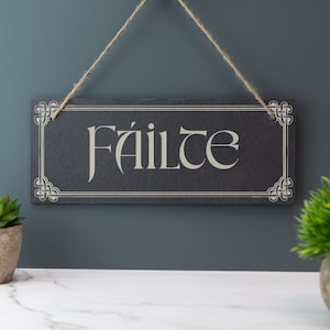 May include: A rectangular black slate sign with the word "Fáilte" in a decorative font. The sign is framed with a white border and corner designs, and is suspended by a natural twine. The sign is hanging against a dark teal wall.