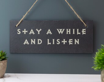 Puede incluir: Un letrero rectangular de pizarra gris oscuro con las palabras "STAY A WHILE AND LISTEN" en letras blancas. El letrero está suspendido por un cordón de hilo natural. El fondo es de color verde azulado apagado.