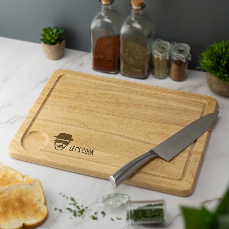 May include: A wooden cutting board with a  'Let's Cook' inscription and a cartoon image of a man wearing a hat and sunglasses. A silver knife is on the board.