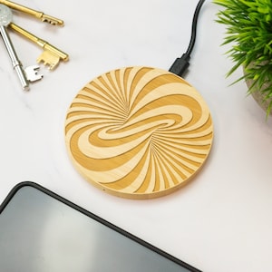 May include: A round, wooden wireless charging pad with a swirling, infinity symbol design. The pad is made of light brown bamboo and has a black USB cable attached.