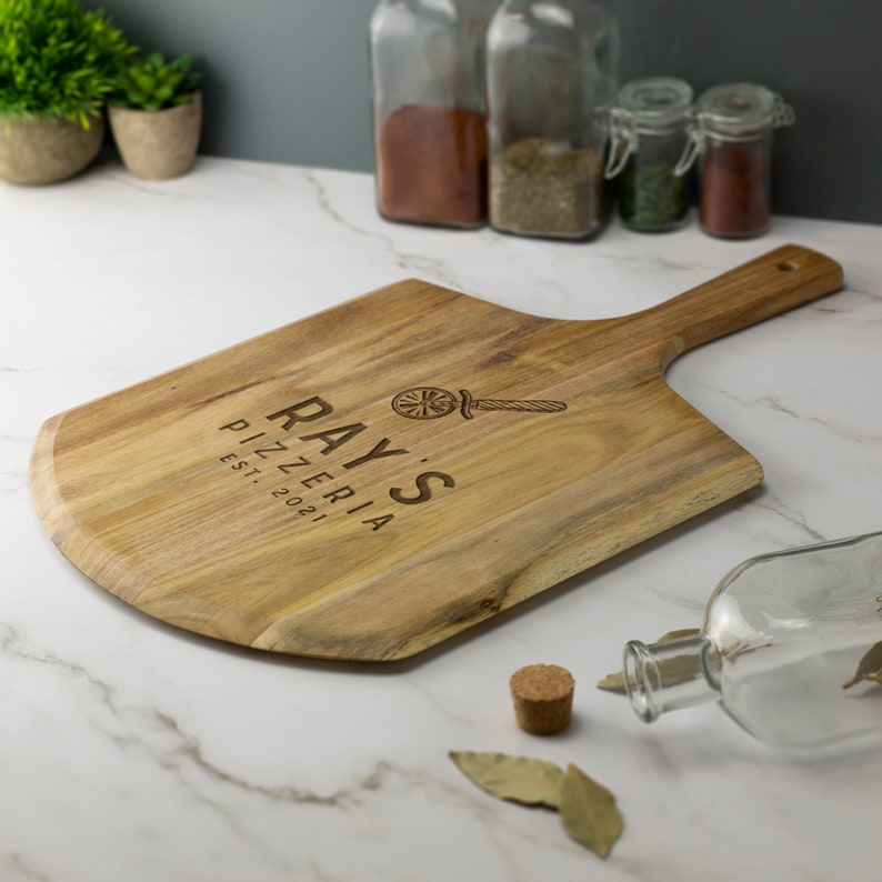 May include: A wooden pizza peel with a handle, engraved with the text "Ray's Pizzeria Est. 2021" and a graphic of a pizza wheel and a pizza cutter.