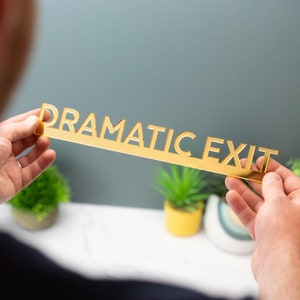 K&ouml;nnte beinhalten: Ein goldfarbenes Acrylschild mit den ausgeschnittenen Worten "Dramatic Exit". Das Schild wird von den H&auml;nden einer Person gehalten.