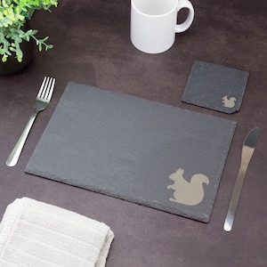 Puede incluir: Una tabla de servir de pizarra gris y un posavasos a juego con un diseño de ardilla. También hay una taza blanca, cubiertos y una toalla blanca doblada. La tabla es rectangular y tiene textura.