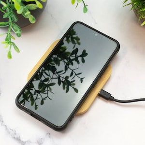 May include: A black smartphone is charging on a wooden wireless charging pad. A black charging cable is connected to the pad.