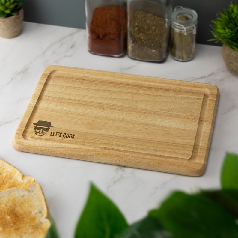May include: A wooden cutting board with a cartoon image of Walter White from the TV show Breaking Bad and the text "Let's Cook".