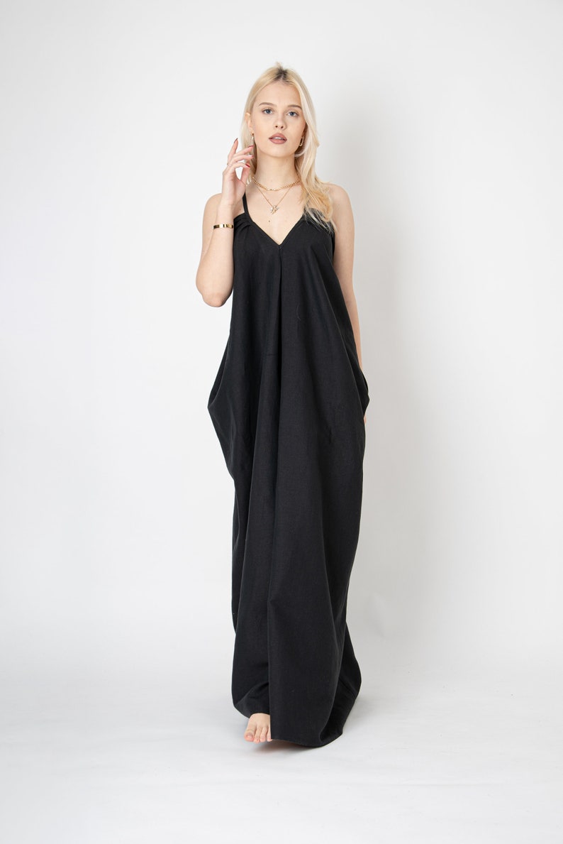 Pode incluir: Uma mulher vestindo um vestido maxi preto com decote em V e al&ccedil;as finas. Ela est&aacute; em p&eacute; em um fundo branco.