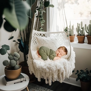 Op de afbeelding: Een pasgeboren baby slaapt in een witte macramé schommelstoel met een wit, pluche deken. De schommel hangt aan het plafond in een kamer met planten.
