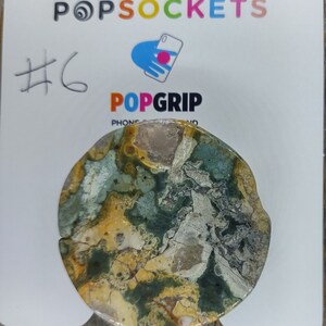 May include: A round, green, yellow, and white patterned phone grip with the text "PopGrip" on the back. The grip is on a white card with the text "#6" and "PopSockets" on it.