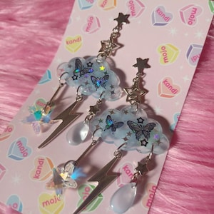 May include: A pair of whimsical earrings featuring a light blue cloud design with butterfly accents. Each earring has a silver-colored lightning bolt, a star, and a teardrop-shaped charm. The earrings are displayed on a pink card with heart-shaped graphics.