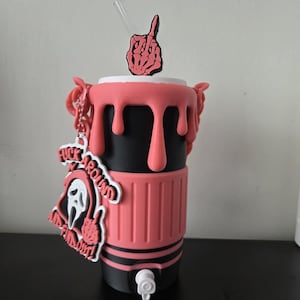 May include: A black and pink tumbler featuring a pink drip design on the rim, a pink middle band, and a white spigot at the base. A skeleton hand middle finger topper sits on the lid, and a charm displays a ghost mask with the text FUCK AROUND AND FIND OUT.