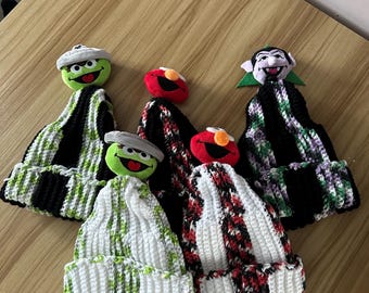May include: A collection of handmade crochet beanies featuring character designs. The hats display a variety of colors, including black, white, green, and red. Each beanie has a character's face attached to the top, such as a green monster, a red character, and a vampire.