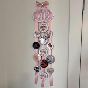May include: A pink button display with a bow at the top, featuring a decorative cloud-shaped element with a monogram. Pink ribbons hold various round buttons with text and graphics, including "War Eagle" and Greek letters.
