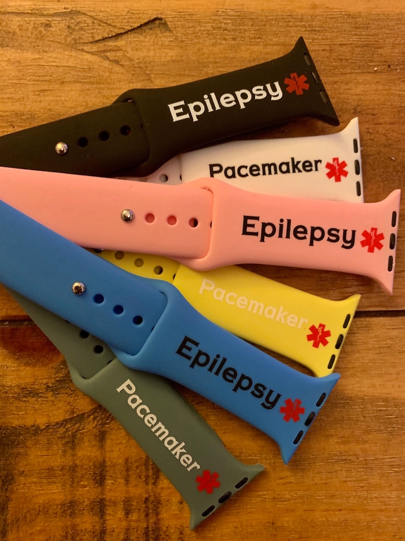 May include: A set of six colorful silicone Apple Watch bands with medical alert text. The bands are black, pink, yellow, blue, and gray. Each band has a red star and the text "Epilepsy" or "Pacemaker".