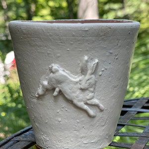 May include: White ceramic planter with a raised design of a rabbit jumping. The planter is sitting on a metal surface.